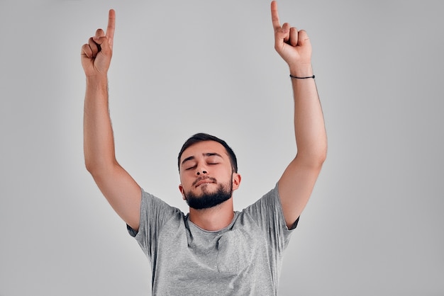 Een jonge man in een grijs T-shirt hief zijn handen op, sloot zijn ogen en genoot van de muziek, overwinning. Studio over grijze achtergrond.