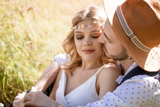 Een jonge man in een hoed en een mooi meisje met een make-up en kapsel, knuffelen zittend in een veld in het gras op een zonnige dag