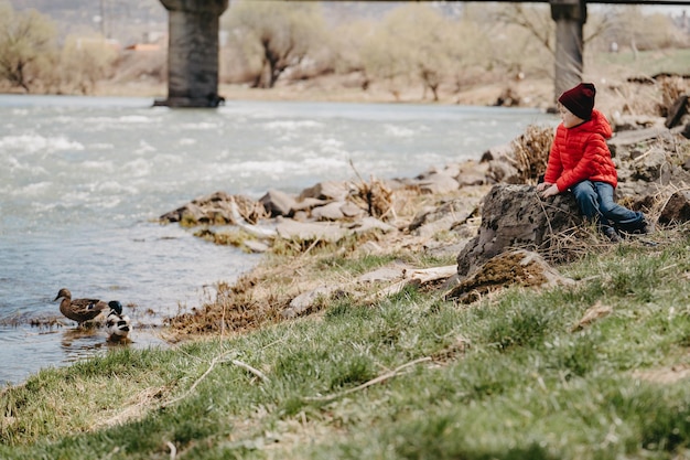 Een jonge man in een rood jasje zit op een rots bij de rivier en kijkt naar de eenden