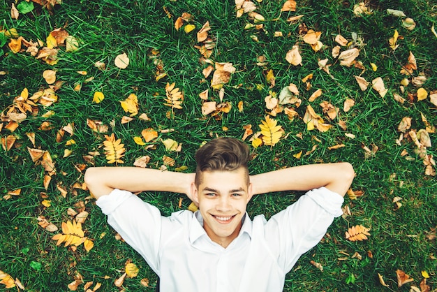 Een jonge man in een wit shirt ligt op het gras met herfstbladeren bovenaanzicht.