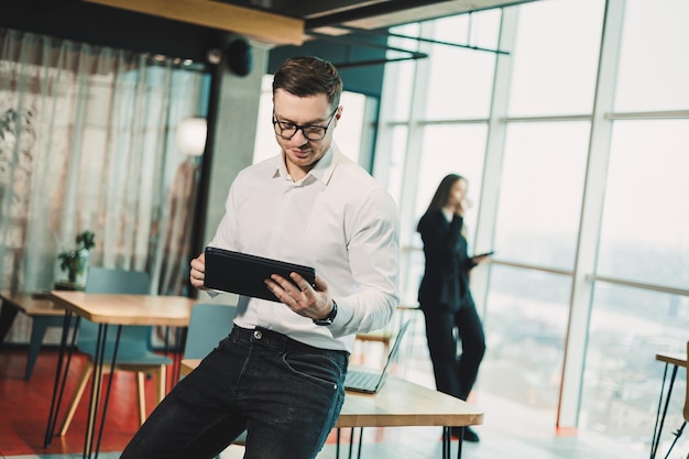 Een jonge man in een wit shirt staat in een ruim modern kantoor en werkt op een tablet Moderne kantoormedewerker Een zakenman in een bril werkt online