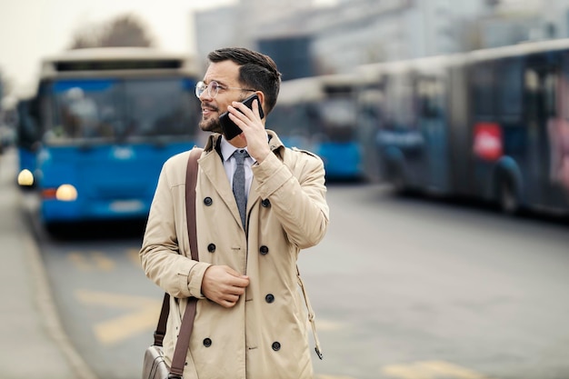Een jonge man in smart casual is aan de telefoon terwijl hij wacht op een openbare bus op een station