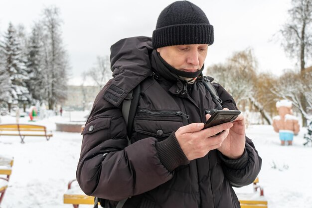 Een jonge man in zwarte winterkleren staat op straat met een telefoon in zijn handen Een man in een warme zwarte jas en hoed kijkt naar zijn telefoon op een winterse achtergrond De stad ligt vol sneeuw