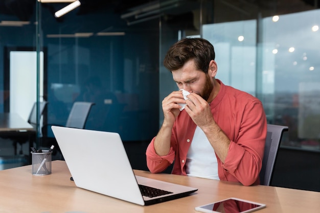 Een jonge man is ziek op het werk, zit aan het bureau op kantoor, veegt zijn neus af met een servet en heeft een loopneus