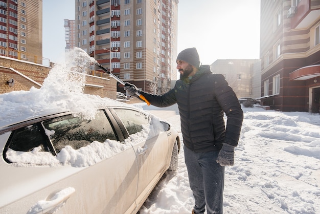 Een jonge man maakt zijn auto schoon na een sneeuwval op een zonnige, ijzige dag.