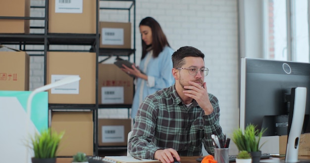 Een jonge man met een bril gebruikt een desktopcomputer een vrouw sorteert artikelen in kartonnen dozen Medewerkers bereiden verkooporders voor in de online winkel