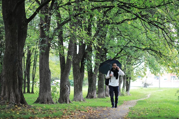 Een jonge man met een bril loopt in het park met een paraplu tijdens de regen.