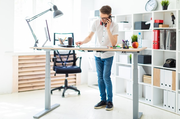 Foto een jonge man met een bril staat bij een computerbureau en praat aan de telefoon. voor hem ligt een magnetisch bord en markers. in de nek hangt de koptelefoon van de man.