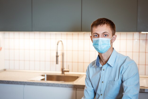 Een jonge man met een kaukasisch uiterlijk zit in een lichtblauw shirt en een medisch masker in de keuken ook...