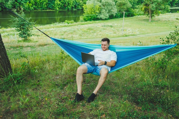 Een jonge man met een laptop zit in een hangmat in de natuur en werkt op afstand Werk in de natuur tijdens de vakantie