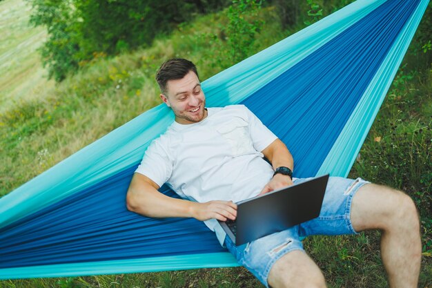 Een jonge man met een laptop zit in een hangmat in de natuur en werkt op afstand. Werk in de natuur tijdens vakantie