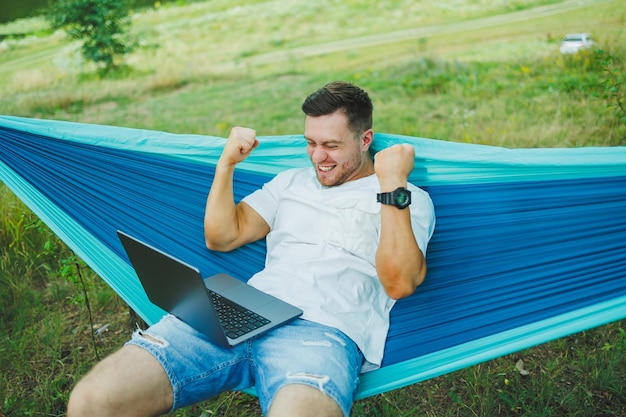 Een jonge man met een laptop zit in een hangmat in de natuur en werkt op afstand. Werk in de natuur tijdens vakantie