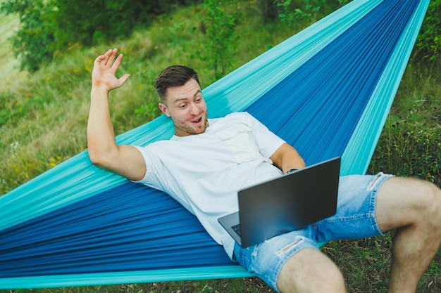 Een jonge man met een laptop zit in een hangmat in de natuur en werkt op afstand. Werk in de natuur tijdens vakantie