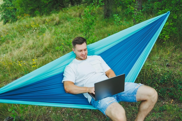 Een jonge man met een laptop zit in een hangmat in de natuur en werkt op afstand. Werk in de natuur tijdens vakantie