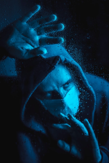 Een jonge man met een masker en een capuchon kijkt uit het raam in de covid19 quarantaine op een regenachtige nacht, met blauw omgevingslicht