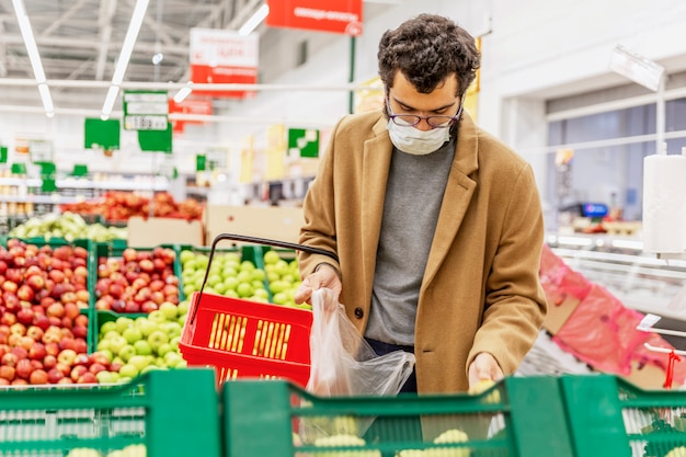 Een jonge man met een medisch masker kiest fruit in een grote supermarkt. Voorzorgsmaatregelen tijdens de pandemie van het coronavirus. Gezond eten.