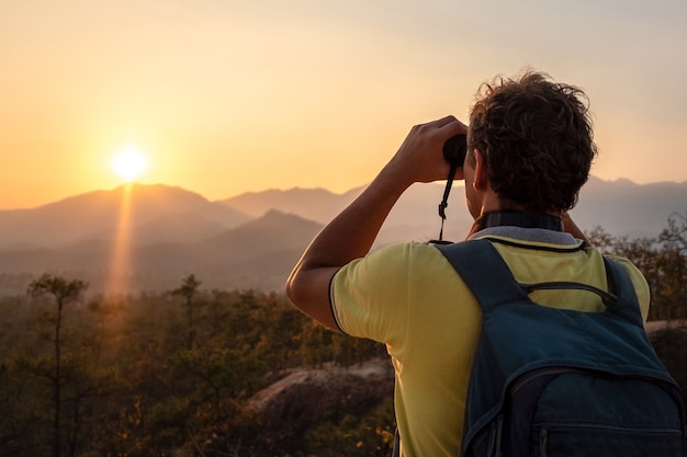 Een jonge man met een rugzak op zijn rug kijkt door een verrekijker naar de zonsondergang naar de silhouetten van de bergen.