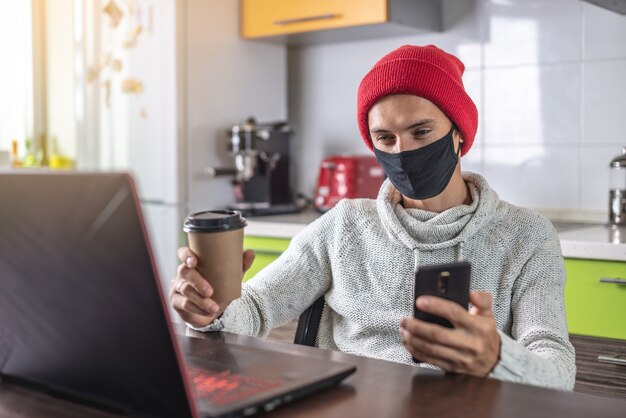 Een jonge man met een zwart gezichtsmasker gebruikt een laptop en een telefoon terwijl hij thuis werkt