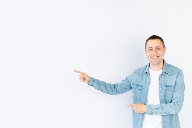Een jonge man of man in een blauw denim overhemd op een witte geïsoleerde lege achtergrond in een fotostudio wijst zijn vinger naar de lege ruimte aan de zijkant voor tekst