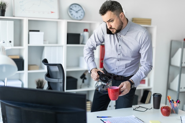 Een jonge man op kantoor staat bij de tafel, houdt de telefoon met zijn schouder vast, kijkt naar de monitor en schenkt koffie in de beker.