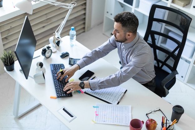 Een jonge man op kantoor zit aan een tafel, houdt een marker in zijn hand en werkt met documenten en een computer.