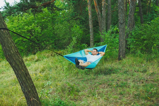 Een jonge man rust in de natuur, zittend in een toeristische hangmat Hangmat voor openluchtrecreatie Weekendrust van een eenzame man in een verlaten bos