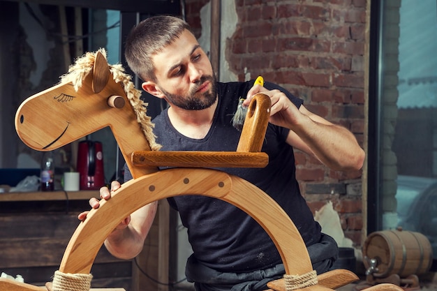 Foto een jonge man schildert een houten speelgoed met een tassel van zijn eigen productie van hout uit de werkplaats
