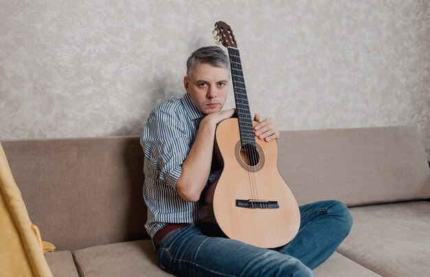 Een jonge man speelt gitaar zittend op een bank in een lichte woonkamer. Een muziekinstrument voor het concept vrije tijd of hobby. Home Entertainment