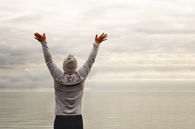 Een jonge man staat aan de kust. Het uitzicht vanaf de achterkant. Yoga lessen. Handen omhoog. Vrijheid en prestatie.