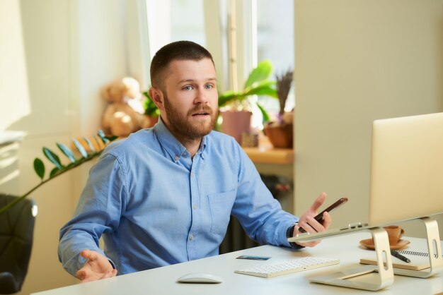 Een jonge man vormt zittend aan een wit bureau voor een laptopcomputer en heeft een smartphone