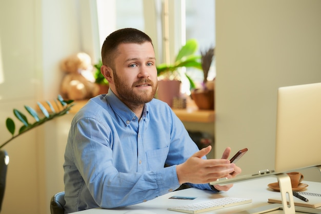 Een jonge man vormt zittend aan een wit bureau voor een laptopcomputer en heeft een smartphone