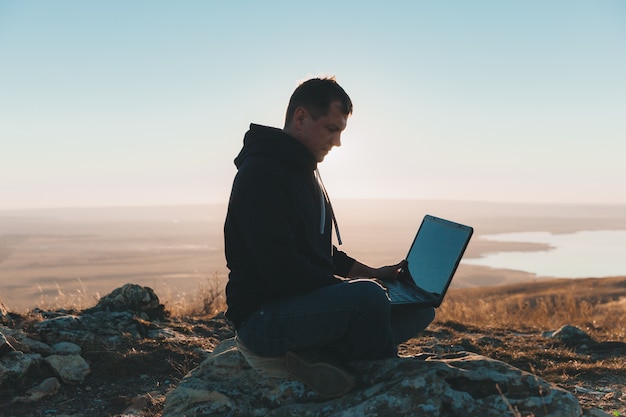 Een jonge man zit op een heuvel en gebruikt een laptop. digitale nomaden.