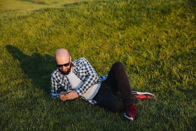 Een jonge man zit op het groene gras in het park en gebruikt sociale netwerken met een mobiele telefoon