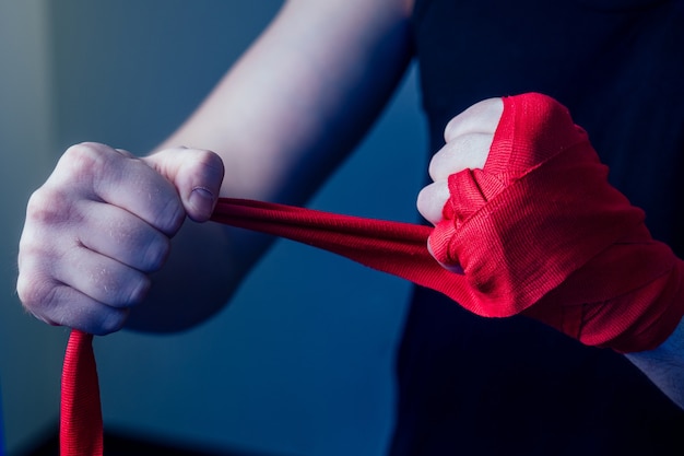 Een jonge mannelijke bokser doet aan sport in de sportschool. bokser, trekt bokshandschoenen aan op een donkere achtergrond. de man slaat toe. rood verband op de handen