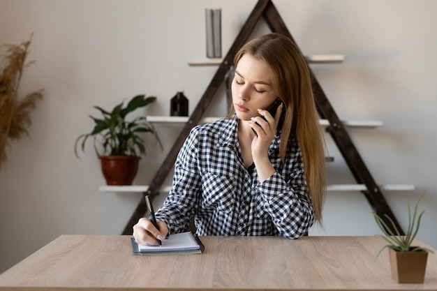 Een jonge meisjesmanager werkt en zit alleen op kantoor aan de tafel, pratend aan de telefoon en schrijvend in een notitieboekje Externe bedrijfs- of onderwijsconcept