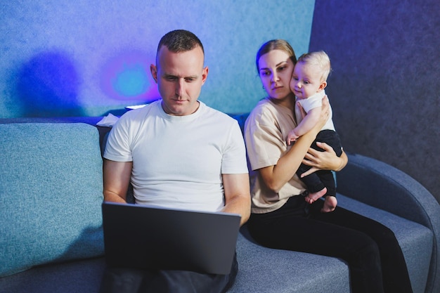 Een jonge moeder en vader zitten met hun pasgeboren zoon op de bank en kijken naar een film op een laptop Gezinsrelaties met een kleine baby Een gezinspaar met een kind thuis op de bank