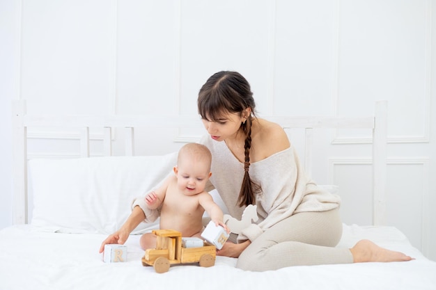 Een jonge moeder speelt met een baby in luiers met houten speelgoed op een licht bed thuis en glimlacht