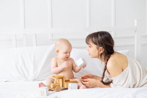 Een jonge moeder speelt met een baby in luiers met houten speelgoed op een licht bed thuis en glimlacht