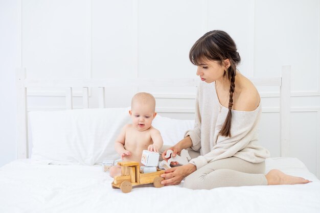 Een jonge moeder speelt met een baby in luiers met houten speelgoed op een licht bed thuis en glimlacht