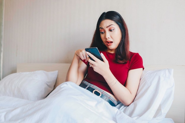 Een jonge, mooie Aziatische vrouw die geschokt op bed zit terwijl ze haar smartphone vasthoudt