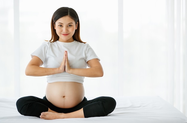 Een jonge mooie Aziatische vrouw zittend op een bed sluit zich aan bij haar handen voor haar borst met een smiley die naar een camera kijkt. Yoga is een geweldige oefening voor zwangere dames.