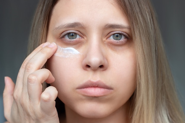 Een jonge mooie blanke blonde vrouw raakt de huid van haar gezicht terwijl ze de crème voor zwarte ogen aanbrengt.