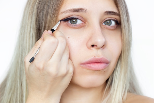 Een jonge mooie blonde vrouw die een contourborstel gebruikt tijdens het tekenen van pijlen op de geïsoleerde oogleden. Een meisje maakt thuis zelf make-up