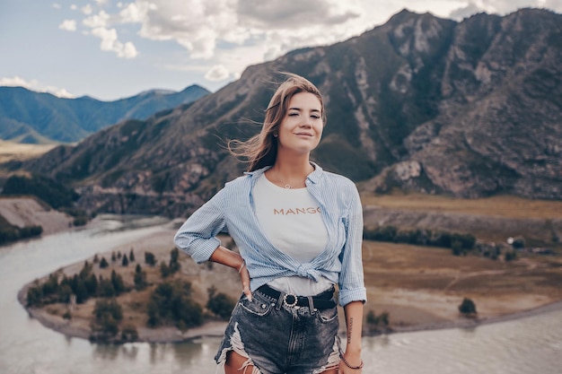 Een jonge mooie brunette in denim shorts en een shirt lacht zoet poseert tegen de achtergrond van rotsachtige bergen en een stromende rivier. Natuurlijke fotosessie.