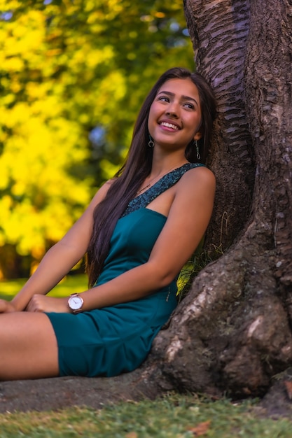 Een jonge mooie brunette Latina met lang steil haar, leunend tegen een boom in een groene jurk.