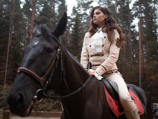 Een jonge mooie brunette ruiter in een elegant retro pak op een zwart paard in een bosgebied