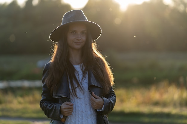 Een jonge, mooie langharige vrouw in een hoed glimlacht, kijkt naar de camera in het park in de heldere gloed van zonsondergangstralen