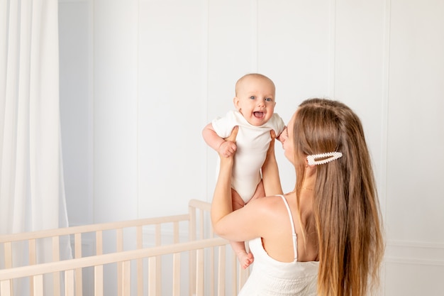 Een jonge, mooie moeder houdt haar dochter van 6 maanden oud in haar armen en tilt haar op in de kinderkamer bij de wieg, Moederdag