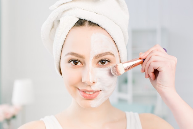 Een jonge mooie vrouw in een badhanddoek na het douchen en het reinigen van de huid brengt een kleimasker aan op het gezicht met een cosmetische borstel. schoonheid, gezondheid, cosmetologieconcept. Levensstijl. Huidverzorging