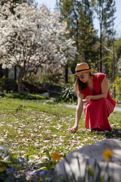 Een jonge, mooie vrouw in een elegante rode jurk en strohoed verzamelt de bloembladen van witte magnoliabloemen die op de grond zijn gevallen in een lentebloeiende tuin